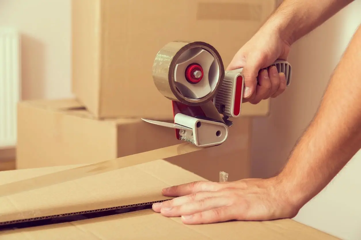 Taping cardboard box with tape dispenser, illustrating packing services by Hilton Moving and Storage.